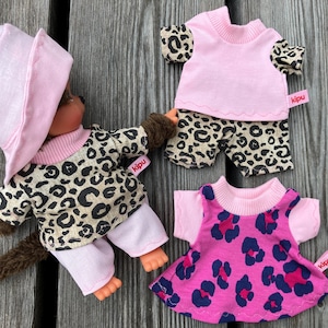 Könnte beinhalten: Eine Puppe mit einem rosa Fischerhut und einem Leopardenmuster-Shirt mit rosa Besatz, kombiniert mit einer hellrosa Hose. Zwei weitere Outfits sind enthalten: ein Leopardenmuster-Shirt mit rosa Weste und ein rosa Kleid mit dunklem Leopardenmuster.