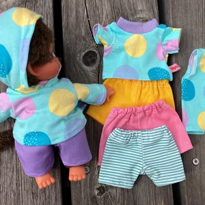 Könnte beinhalten: Eine braun behaarte Puppe in einem hellblauen Kapuzenpullover mit gelben, rosa und blauen Punkten und einer violetten Hose, dazu ein Set Puppenkleidung mit passendem Oberteil, gelber Shorts, rosa Shorts und einer gestreiften Shorts.