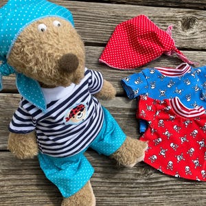 Könnte beinhalten: Ein brauner Teddybär, der ein blau-weiß gestreiftes Hemd, eine blau-weiß gepunktete Hose und ein blau-weiß gepunktetes Bandana trägt. Der Bär hält ein rot-weißes Bandana mit weißen Punkten. Der Bär trägt auch ein rot-weißes Hemd mit Totenkopf- und Knochenmuster.