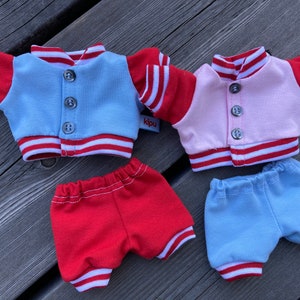 May include: Four miniature doll outfits. Two varsity jackets, one light blue and one pink, with red sleeves and white stripes. Two pairs of shorts, one red and one light blue, with white stripes. The jackets have three buttons.