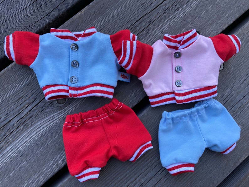 May include: Four miniature doll outfits. Two varsity jackets, one light blue and one pink, with red sleeves and white stripes. Two pairs of shorts, one red and one light blue, with white stripes. The jackets have three buttons.