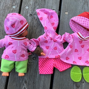 May include: Three pink doll outfits with a lollipop and heart print. One outfit is a hooded top and shorts, another is a dress, and the third is a long-sleeved shirt and trousers. The outfits are all pink with white hearts and lollipops. The dress has a white trim around the neck and sleeves. The trousers are green. The outfits are all made by the brand "Kipu".