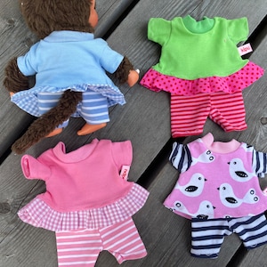 May include: Four doll outfits, including a blue and white striped outfit, a pink and white striped outfit, a green and pink polka dot outfit, and a pink and white bird print outfit. Each outfit has a ruffled top and a pair of pants. The outfits are all made by the brand "Haba".
