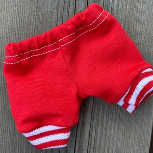 May include: Red shorts with white striped cuffs and waistband. The shorts are made of a soft, knit fabric and feature white stitching details. The cuffs have a classic athletic design.