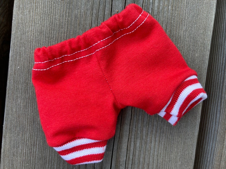 May include: Red shorts with white striped cuffs and waistband. The shorts are made of a soft, knit fabric and feature white stitching details. The cuffs have a classic athletic design.