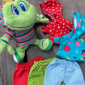 Könnte beinhalten: Ein grünes Frosch-Plüschtier, das ein gestreiftes Shirt trägt. Der Frosch ist von vier Kleidungsstücken umgeben: einem roten Kapuzenpullover mit weißen Punkten, einem blauen Kapuzenpullover mit rosa und roten Punkten, einer roten Hose mit weißen Punkten und einer blauen Hose mit weißen Punkten.