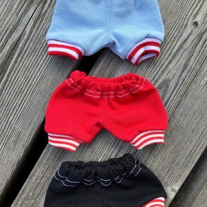 May include: Three pairs of doll-sized shorts in blue, red, and black. Each pair has a contrasting red and white striped trim at the leg openings and a gathered waistband. The shorts are made of a soft fabric and are arranged on a wooden surface.