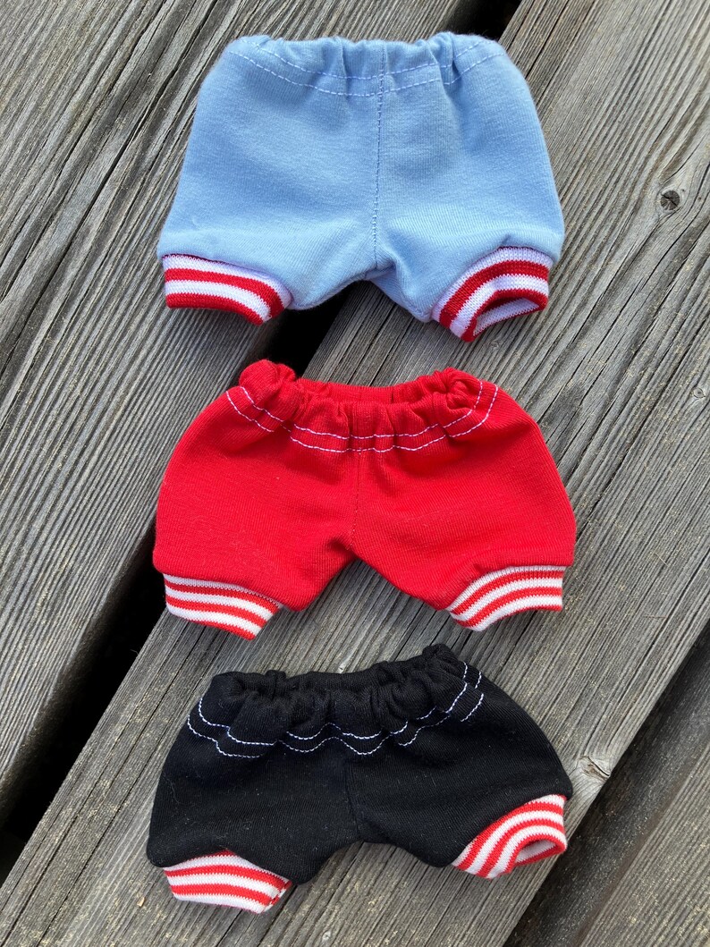 May include: Three pairs of doll-sized shorts in blue, red, and black. Each pair has a contrasting red and white striped trim at the leg openings and a gathered waistband. The shorts are made of a soft fabric and are arranged on a wooden surface.