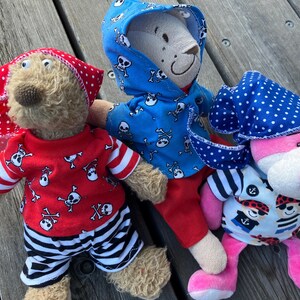 May include: Three plush teddy bears wearing pirate-themed outfits. One bear is wearing a blue hooded shirt with a skull and crossbones pattern, another is wearing a red shirt with a skull and crossbones pattern and black and white striped trousers, and the third bear is wearing a blue and white polka dot bandana and a blue and white striped shirt with a pirate graphic.