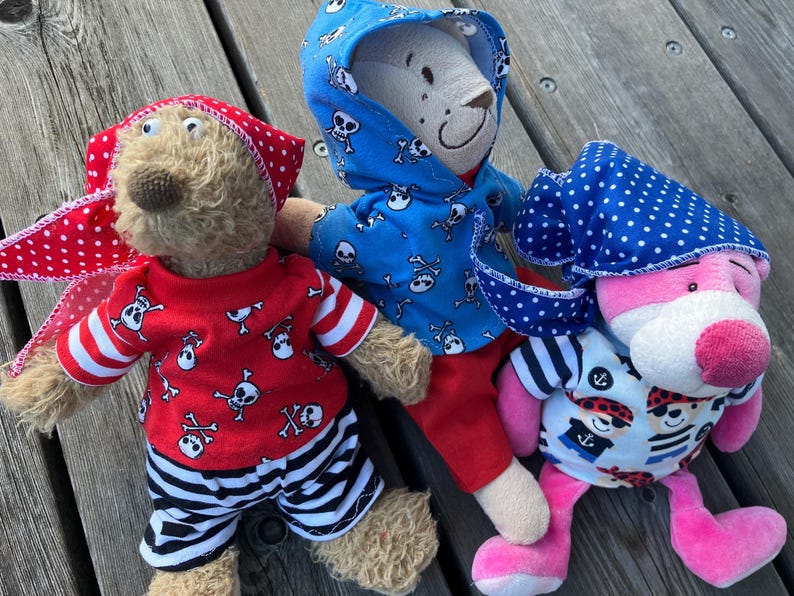 May include: Three plush teddy bears wearing pirate-themed outfits. One bear is wearing a blue hooded shirt with a skull and crossbones pattern, another is wearing a red shirt with a skull and crossbones pattern and black and white striped trousers, and the third bear is wearing a blue and white polka dot bandana and a blue and white striped shirt with a pirate graphic.