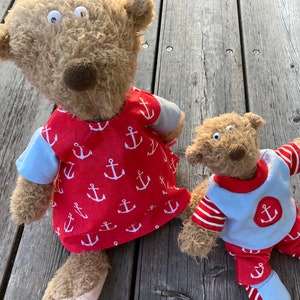 Könnte beinhalten: Zwei braune Teddybären, die rote und blaue Kleidung mit weißen Ankermustern tragen. Der größere Bär trägt ein rotes Kleid mit weißen Ankern und ein blaues Kurzarmhemd. Der kleinere Bär trägt ein blaues Hemd mit einem roten Anker und rot-weiß gestreifte Hosen.
