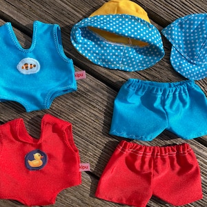 Könnte beinhalten: Zwei Sets Puppenkleidung für eine Puppe. Ein Set ist blau mit einem Fischdesign und das andere ist rot mit einem Gummiente-Design. Es gibt auch zwei blau-weiße gepunktete Hüte.