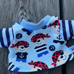 May include: A light blue t-shirt featuring a pattern of bears wearing pirate hats and eyepatches. The bears are wearing striped shirts and holding anchors. The shirt has black and white striped sleeves and a blue collar. The shirt has a red label with the word "kipu" on it.