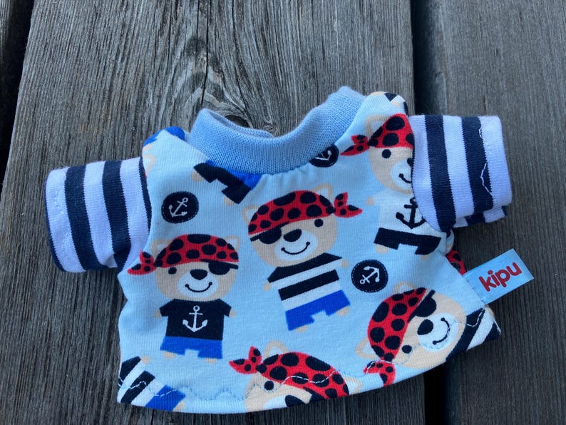 May include: A light blue t-shirt featuring a pattern of bears wearing pirate hats and eyepatches. The bears are wearing striped shirts and holding anchors. The shirt has black and white striped sleeves and a blue collar. The shirt has a red label with the word "kipu" on it.