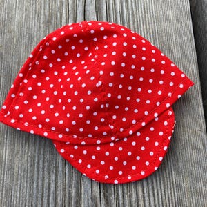 May include: A red cap with a white polka dot pattern. The cap has a curved brim and is made of a fabric material. The cap is laying on a wooden surface.