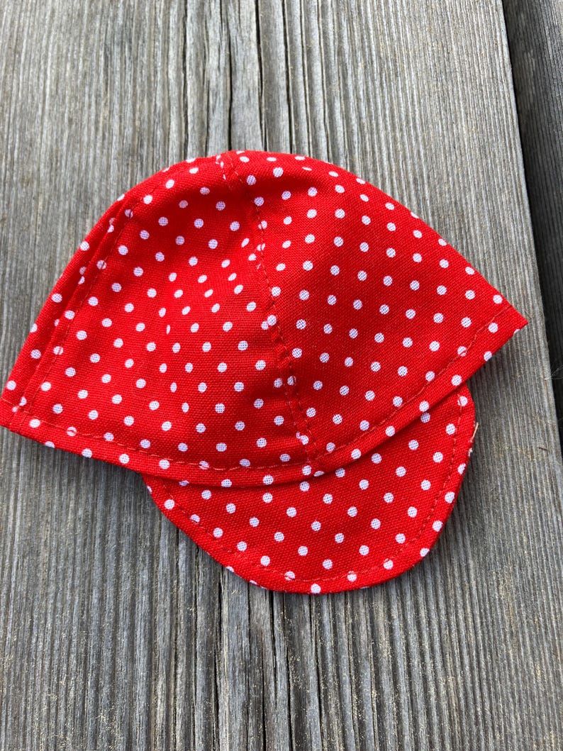 May include: A red cap with a white polka dot pattern. The cap has a curved brim and is made of a fabric material. The cap is laying on a wooden surface.