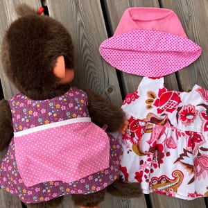 May include: A brown plush monkey doll wearing a purple floral dress with a pink polka dot apron. A pink hat and a white dress with red floral patterns are also displayed. The items are on a wooden surface.