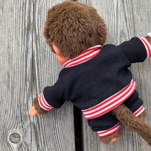 May include: A vintage Monchhichi doll, viewed from the back, wearing a black jacket and pants with red and white striped trim. The doll has brown fur and a tail, resting on a weathered wooden surface.