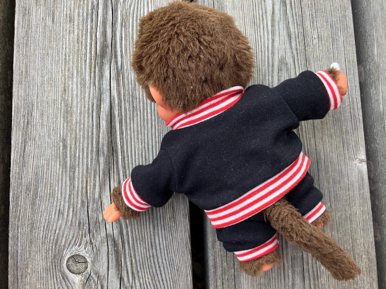 May include: A vintage Monchhichi doll, viewed from the back, wearing a black jacket and pants with red and white striped trim. The doll has brown fur and a tail, resting on a weathered wooden surface.