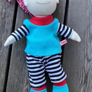 May include: A handmade doll with pink yarn hair, wearing a turquoise shirt with black and white stripes, black and white striped pants, and turquoise shoes. The doll has a red collar and a red trim on the shoes. The doll has a label that says "kipu".
