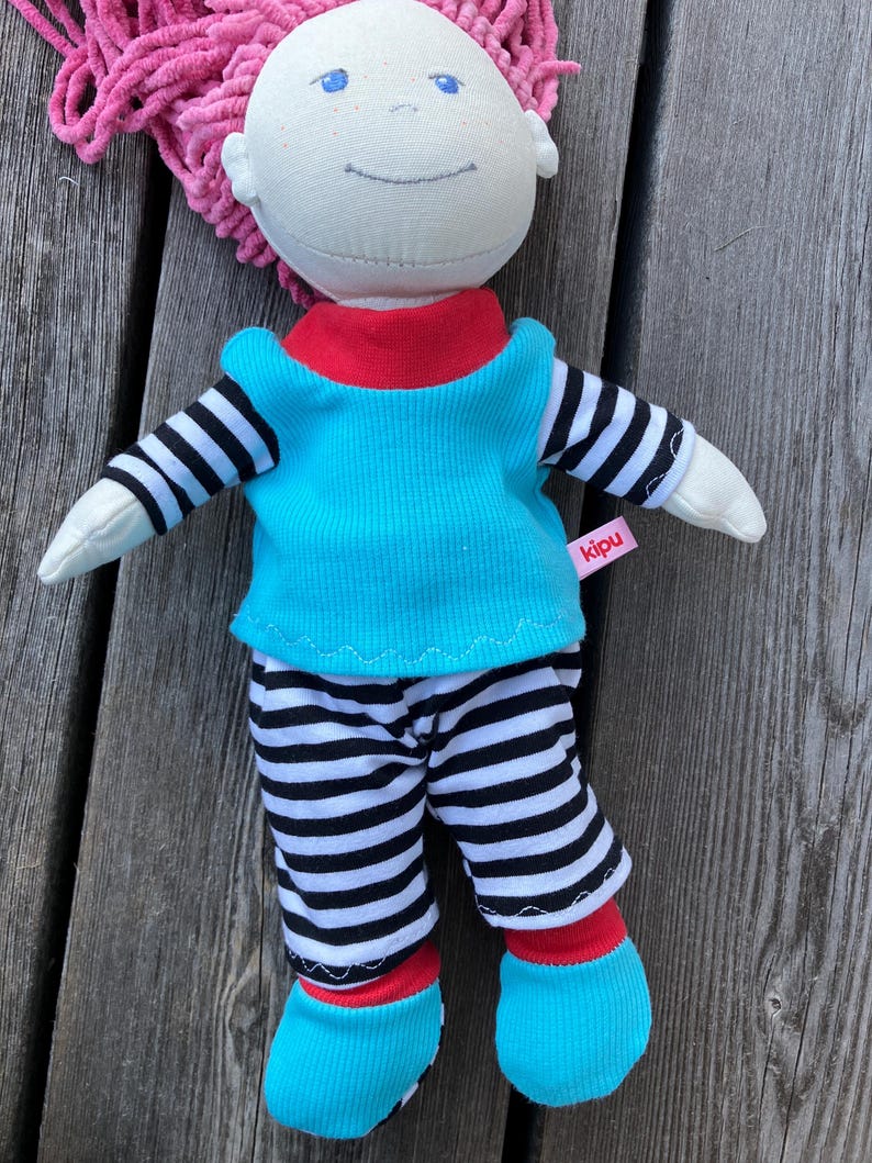 May include: A handmade doll with pink yarn hair, wearing a turquoise shirt with black and white stripes, black and white striped pants, and turquoise shoes. The doll has a red collar and a red trim on the shoes. The doll has a label that says "kipu".