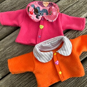 May include: Two fleece doll jackets. One is bright pink with a patterned collar and a heart-shaped button. The other is orange with a striped collar and a heart-shaped button. Both have button closures.