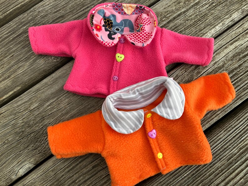 May include: Two fleece doll jackets. One is bright pink with a patterned collar and a heart-shaped button. The other is orange with a striped collar and a heart-shaped button. Both have button closures.