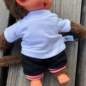 May include: A brown monkey doll wearing a white t-shirt, black shorts with red and white striped trim, and brown fur-like arms and legs. The doll has a tag that says "kipu". The doll is sitting on a wooden surface.