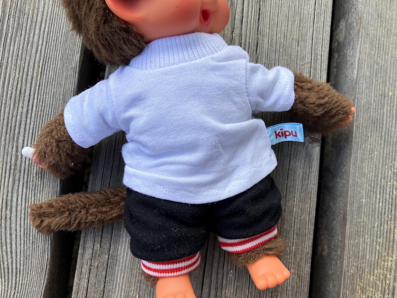 May include: A brown monkey doll wearing a white t-shirt, black shorts with red and white striped trim, and brown fur-like arms and legs. The doll has a tag that says "kipu". The doll is sitting on a wooden surface.