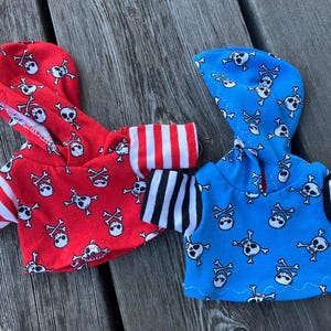 May include: Two hooded tops with skull and crossbones patterns. One top is red with white and black stripes on the sleeves. The other top is blue with black and white stripes on the sleeves. The blue top has a "kipu" tag.