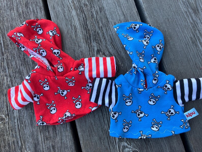 May include: Two hooded tops with skull and crossbones patterns. One top is red with white and black stripes on the sleeves. The other top is blue with black and white stripes on the sleeves. The blue top has a "kipu" tag.