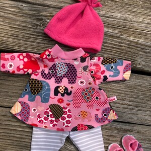 May include: A pink doll outfit set featuring a top with elephant print, striped pants, a pink fleece hat, and matching booties. The top has a pink collar and the pants are gray and white striped.