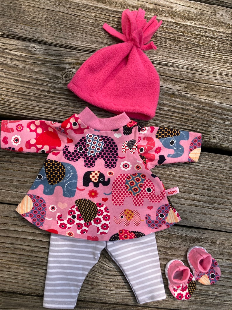May include: A pink doll outfit set featuring a top with elephant print, striped pants, a pink fleece hat, and matching booties. The top has a pink collar and the pants are gray and white striped.