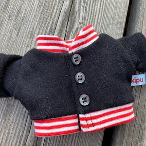 May include: A miniature black jacket with red and white striped trim on the collar, cuffs, and hem. The jacket has three small, clear buttons down the front and a small blue tag that reads "kipu".