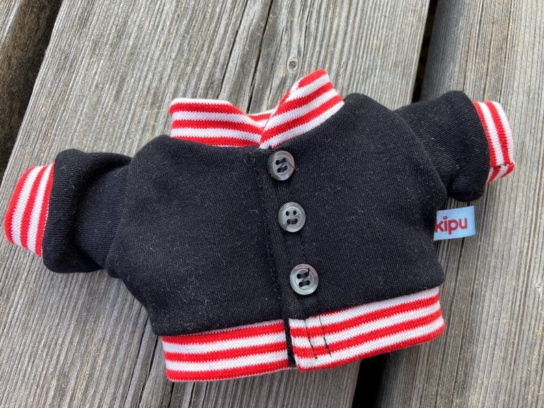 May include: A miniature black jacket with red and white striped trim on the collar, cuffs, and hem. The jacket has three small, clear buttons down the front and a small blue tag that reads "kipu".