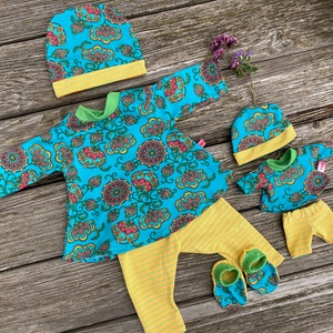 Könnte beinhalten: Ein Baby-Kleidungsset mit einem türkisfarbenen Oberteil und Hut mit floralem Muster, kombiniert mit gelb gestreiften Hosen und passenden Babyschuhen. Ein kleineres Set für eine Puppe ist ebenfalls enthalten. Die Outfits haben grüne und gelbe Akzente.