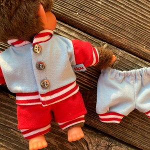 May include: A doll wearing a light blue and red outfit. The jacket has three silver buttons and the word "kipu" on a tag. The shorts are light blue with red and white trim. The doll has brown fur.