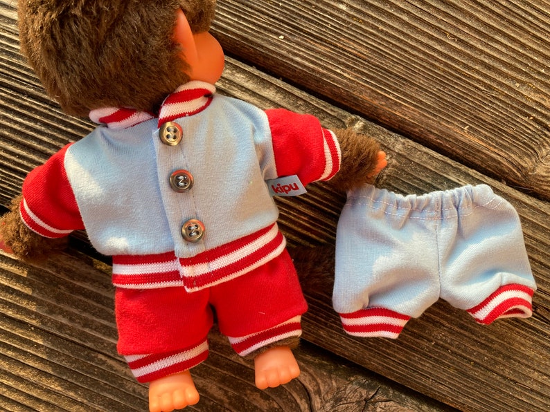 May include: A doll wearing a light blue and red outfit. The jacket has three silver buttons and the word "kipu" on a tag. The shorts are light blue with red and white trim. The doll has brown fur.