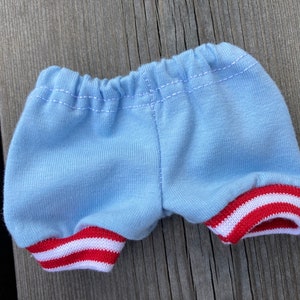 May include: Light blue shorts with red and white striped trim on the leg openings. The shorts have an elastic waistband and are made of a soft fabric. The shorts are designed for a doll or small toy.