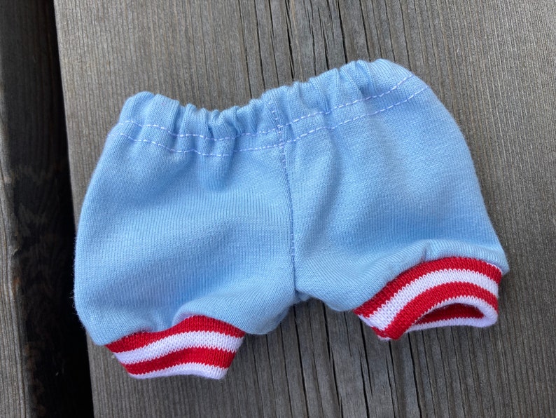 May include: Light blue shorts with red and white striped trim on the leg openings. The shorts have an elastic waistband and are made of a soft fabric. The shorts are designed for a doll or small toy.
