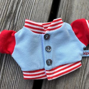 May include: A small, light blue and red varsity jacket with white stripes. The jacket has red sleeves and collar, with white stripes on the cuffs and collar. It has three buttons down the front and a small tag that says "kipu".