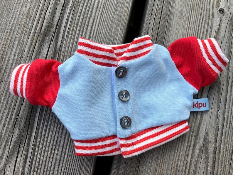 May include: A small, light blue and red varsity jacket with white stripes. The jacket has red sleeves and collar, with white stripes on the cuffs and collar. It has three buttons down the front and a small tag that says "kipu".