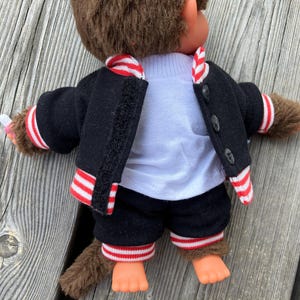 May include: A brown monkey doll wearing a black jacket with red and white striped trim, a white shirt, and black shorts with matching trim. The doll is posed on a weathered wooden surface.