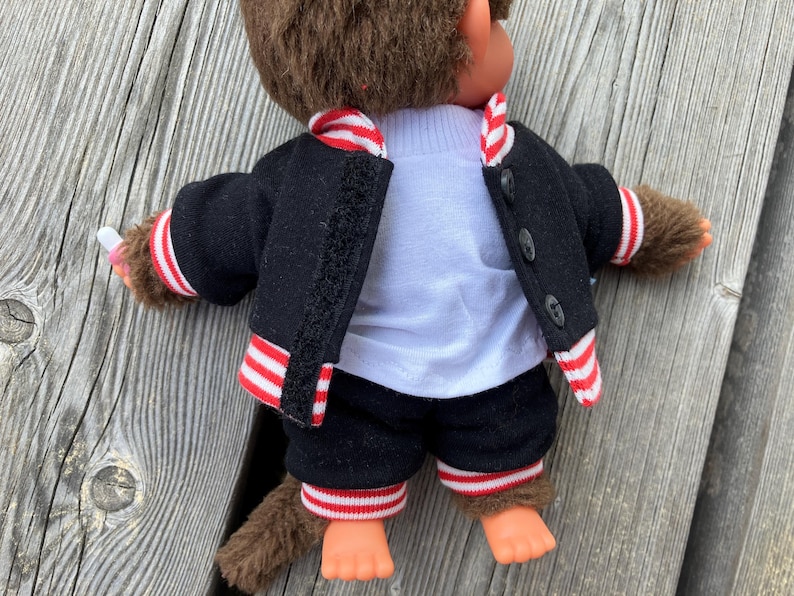May include: A brown monkey doll wearing a black jacket with red and white striped trim, a white shirt, and black shorts with matching trim. The doll is posed on a weathered wooden surface.
