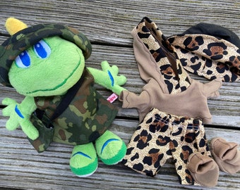 May include: A green plush frog toy wearing a camouflage hat and vest with a small matching bag, alongside a separate outfit consisting of a tan hoodie with a leopard print hood, leopard print shorts, a leopard print bucket hat, and tan booties.
