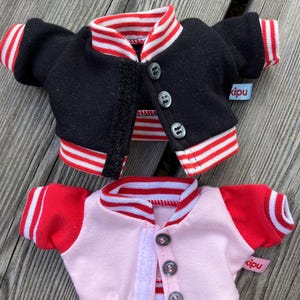 May include: Two miniature varsity jackets, one black and one pink, with red and white striped trim on the collar, cuffs, and hem. Each jacket has a button closure and a small tag that says "kipu".