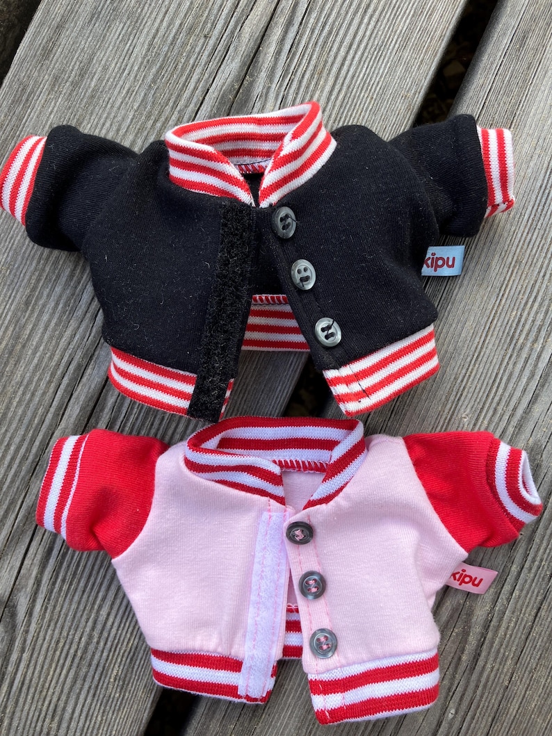 May include: Two miniature varsity jackets, one black and one pink, with red and white striped trim on the collar, cuffs, and hem. Each jacket has a button closure and a small tag that says "kipu".