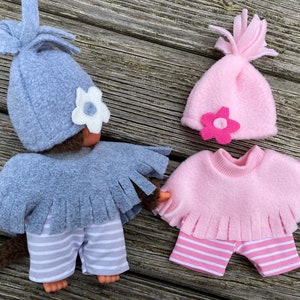Puede incluir: Dos muñecas de peluche de mono que llevan atuendos a juego. La muñeca de la izquierda lleva un gorro gris con una flor blanca, un poncho gris con flecos y pantalones a rayas grises y blancas. La muñeca de la derecha lleva un gorro rosa con una flor rosa, un poncho rosa con flecos y pantalones a rayas rosas y blancas. Ambas muñecas tienen pelo marrón.