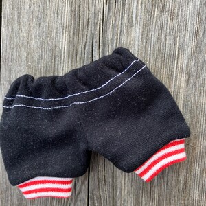 May include: Black shorts with white stitching and red and white striped cuffs. The shorts are made of a soft fabric and appear to be for a doll or small toy. The shorts are displayed on a weathered wooden surface.