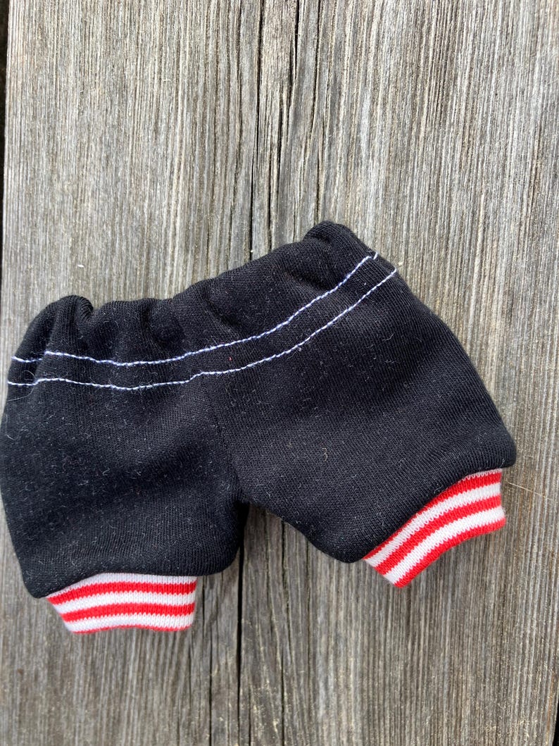 May include: Black shorts with white stitching and red and white striped cuffs. The shorts are made of a soft fabric and appear to be for a doll or small toy. The shorts are displayed on a weathered wooden surface.